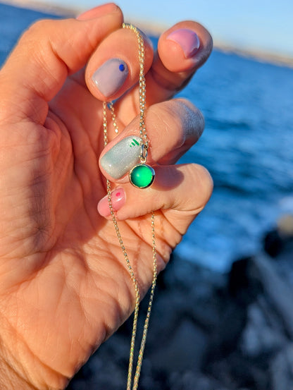 A close-up of a hand holding a delicate gold chain necklace with a green sea glass set in a gold bezel against blurred ocean background.