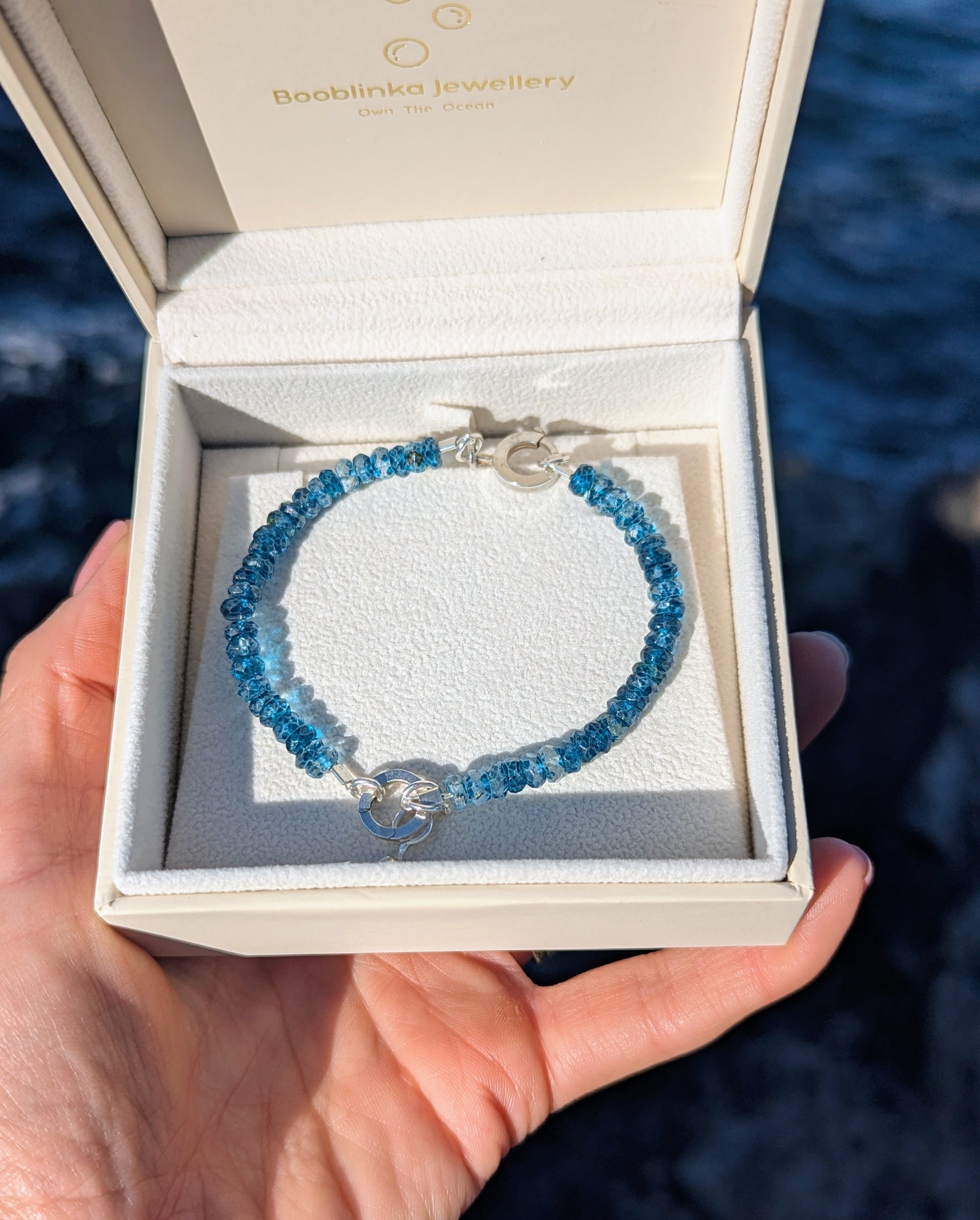A London blue beaded bracelet with a silver clasp and droplet charm, displayed inside an open cream-coloured Booblinka Jewellery gift box held by a hand against a dark blue water background.