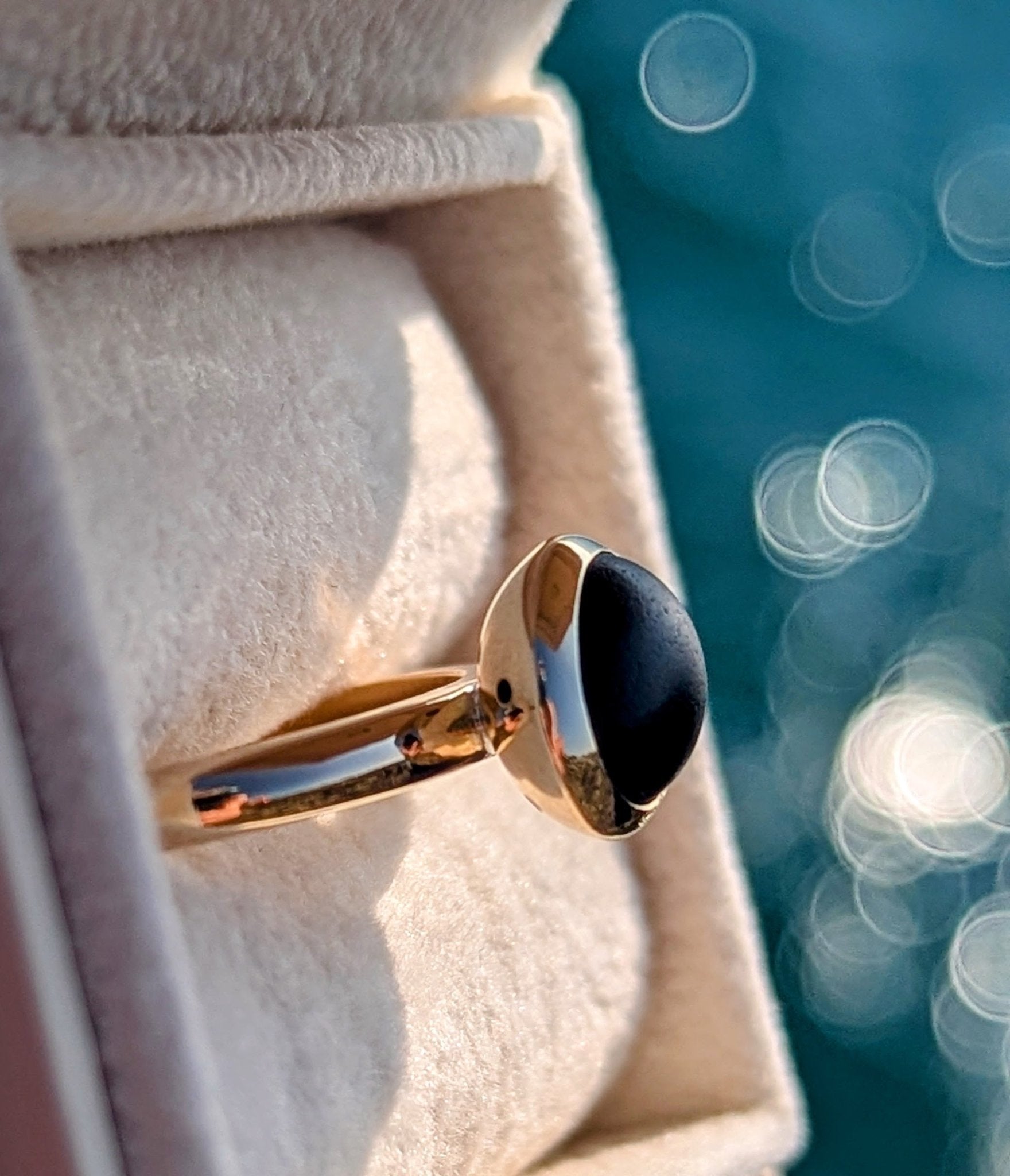 Side view of opaque black sea glass set in a handmade solid 9ct gold ring with blurred ocean sparkling background