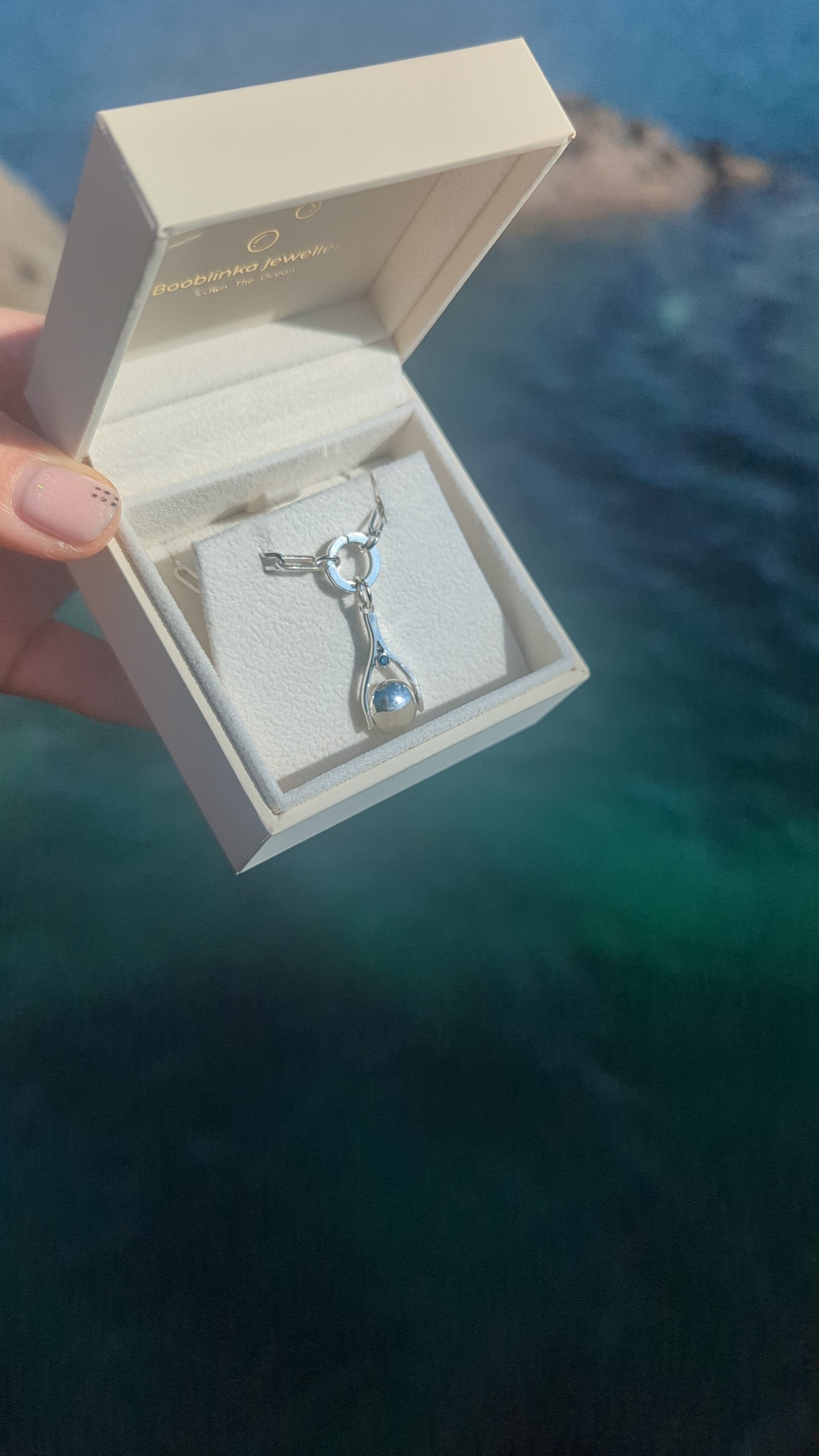 Silver mini spinning minstrel necklace with blue diamonds made by Booblinka Jewellery in a jewellery box above the ocean in Cornwall