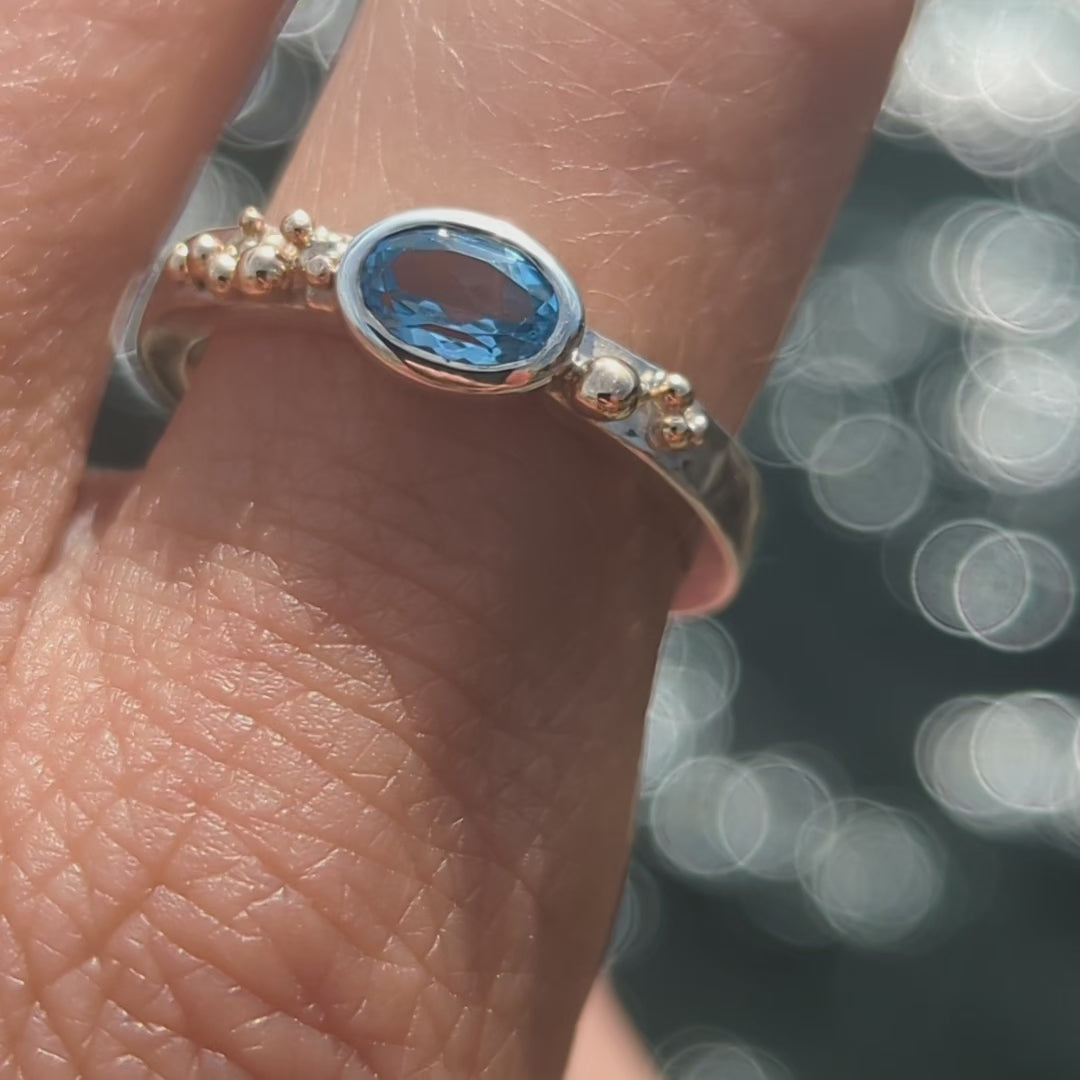 Detail of handmade silver Ring with a Swiss blue oval topaz and gold granules on a finger above shimmering ocean made by Booblinka Jewellery