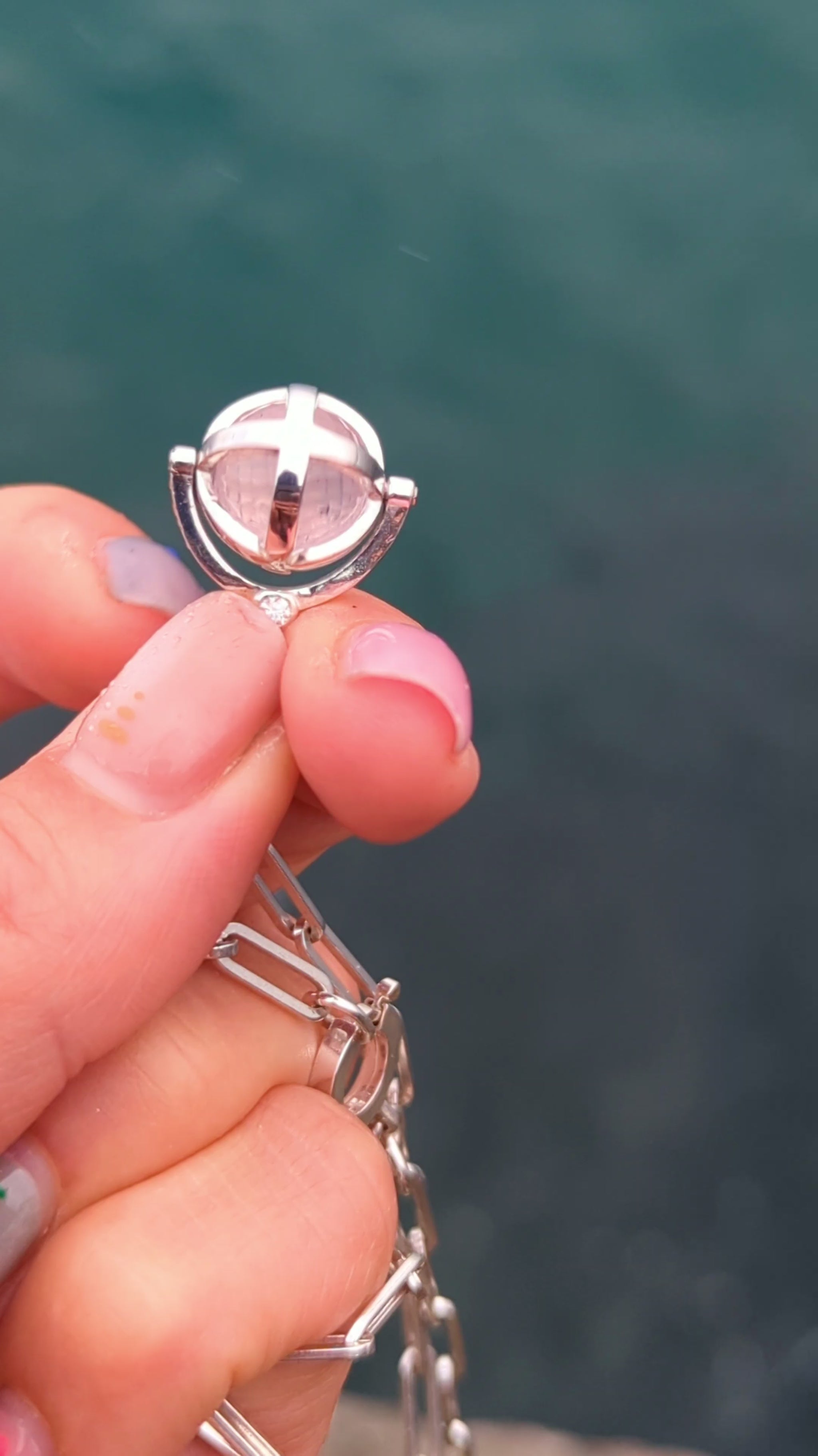 Silver ORB necklace held in a hand while the cage containing the rose quartz sphere is turned and spun by hand.