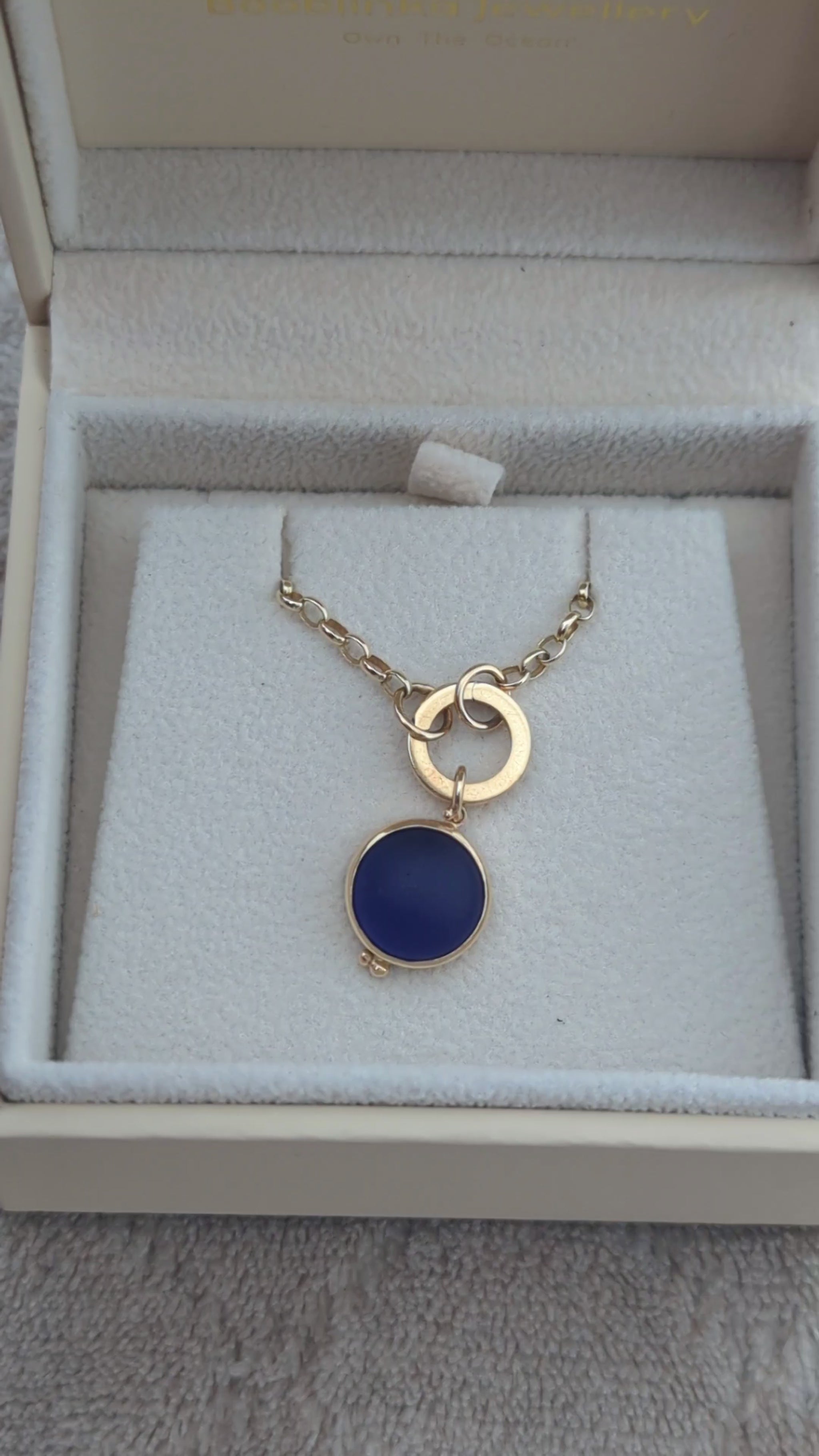 Video showing a round blue sea glass pendant in solid 9ct gold with a hinged gold charm holder and gold chain in a jewellery box.
