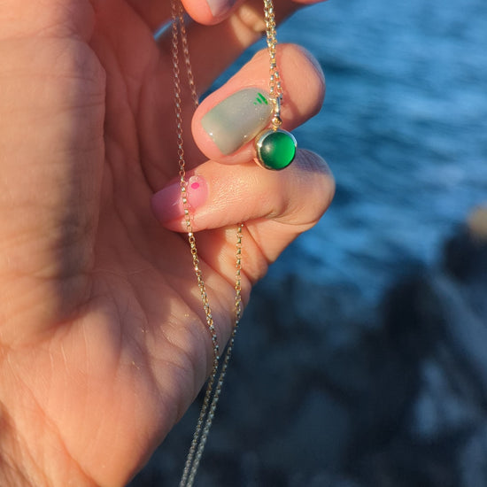 A close-up of a hand holding a delicate gold chain necklace with a green sea glass set in a gold bezel against blurred ocean background.
