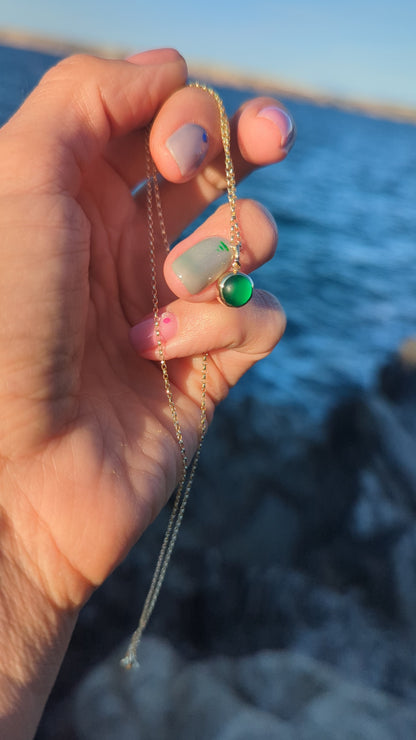 A close-up of a hand holding a delicate gold chain necklace with a green sea glass set in a gold bezel against blurred ocean background.