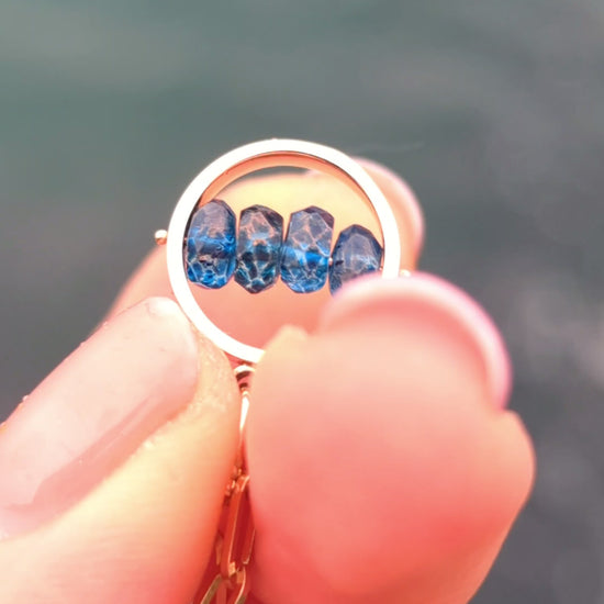 Video of fingers spinning Solid 9ct gold HALO necklace with four faceted blue topaz gemstones across the centre, against the sea.
