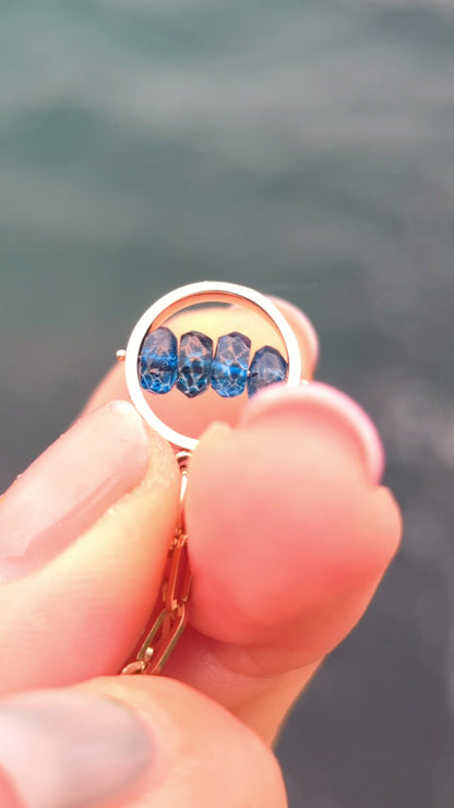 Video of fingers spinning Solid 9ct gold HALO necklace with four faceted blue topaz gemstones across the centre, against the sea.