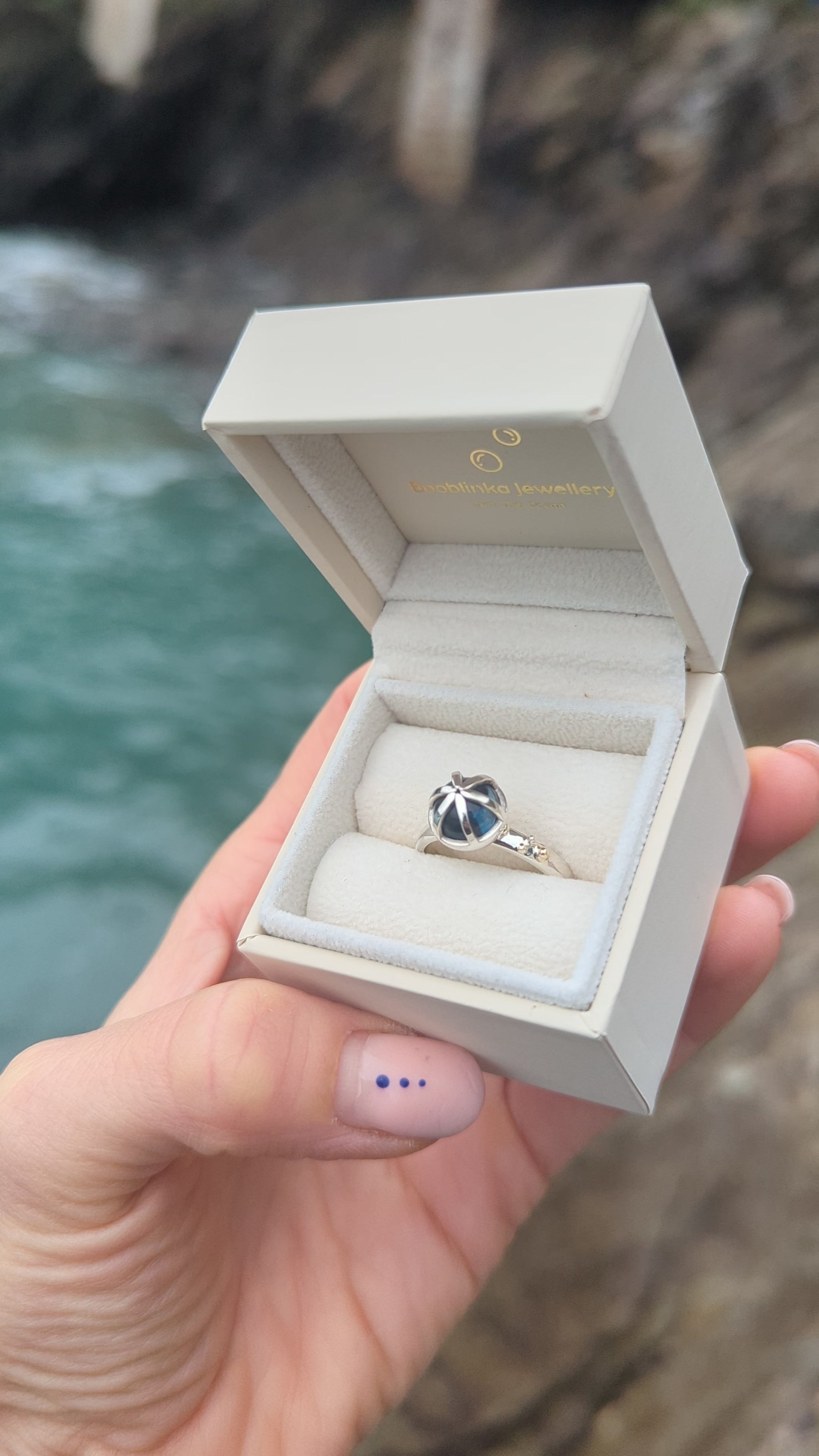 Video of statement silver ring with London blue topaz and gold granules in a jewellery box held in hand above the ocean made by Booblinka Jewellery