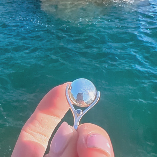 Video of silver spinning necklace with blue topaz spinning above the ocean made by Booblinka Jewellery.