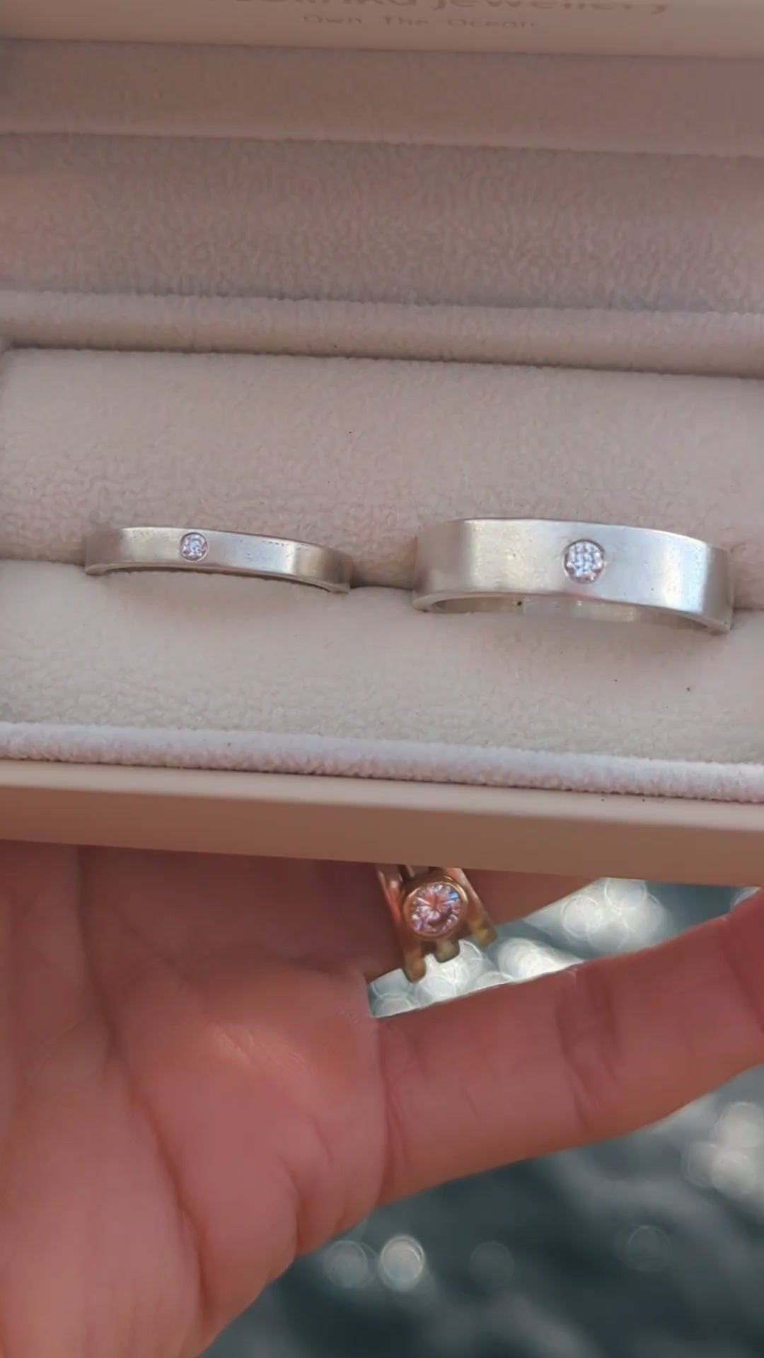 Video showing the two sterling silver wedding bands with flush-set moissanites in an open jewellery box with blurred ocean background.