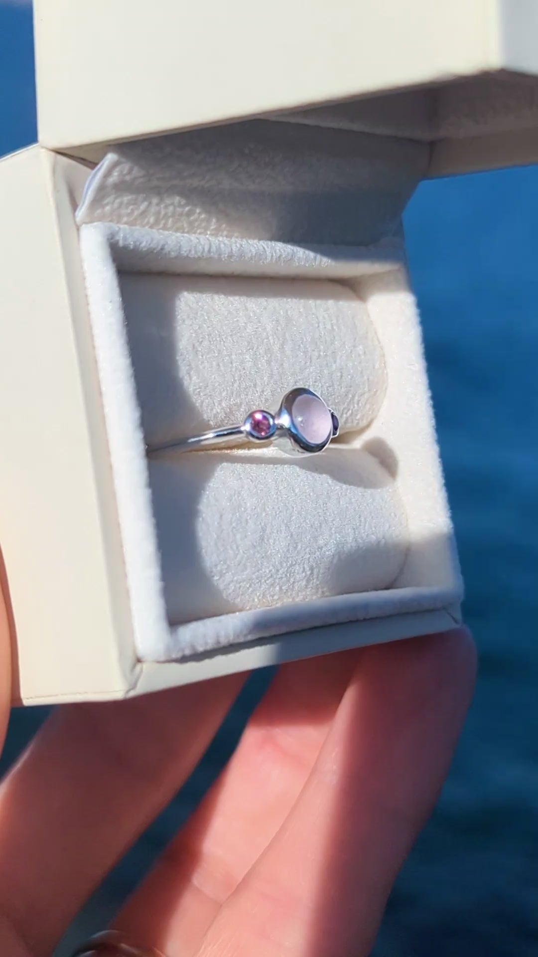 Silver ring with pink sea glass and pink tourmaline gemstones in an ivory jewellery box against a blue ocean made by Booblinka Jewellery