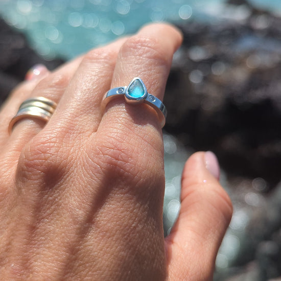 Aqua Blue Sea Glass and Moissanite Silver Ring on a finger above the shimmering ocean
