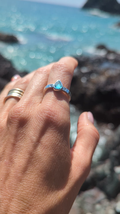 Aqua Blue Sea Glass and Moissanite Silver Ring on a finger above the shimmering ocean