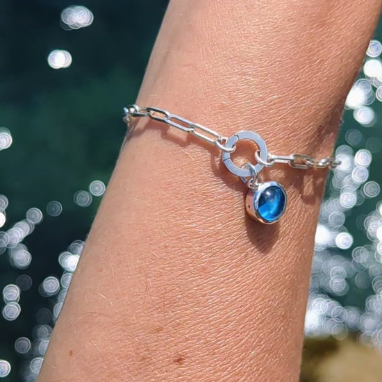 Video showing Silver paperclip bracelet with a Swiss blue topaz charm on a hand against a shimmery water background made by Booblinka Jewellery.