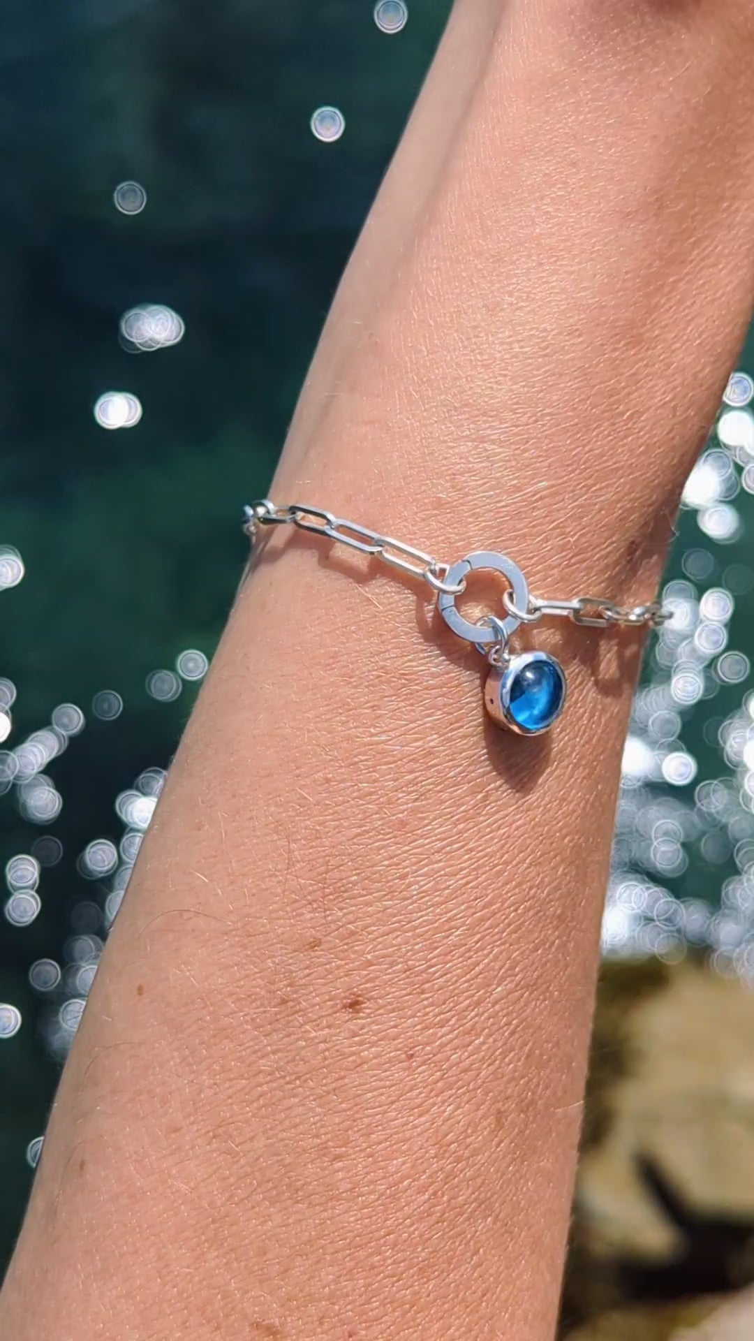Video showing Silver paperclip bracelet with a Swiss blue topaz charm on a hand against a shimmery water background made by Booblinka Jewellery.