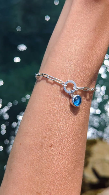 Video showing Silver paperclip bracelet with a Swiss blue topaz charm on a hand against a shimmery water background made by Booblinka Jewellery.