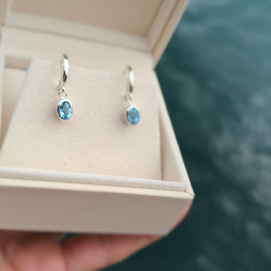 video of  oval Blue topaz huggie silver earrings in an ivory plush jewellery box above the ocean made by Booblinka Jewellery
