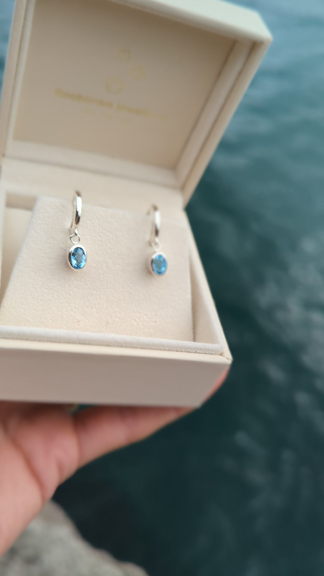 video of  oval Blue topaz huggie silver earrings in an ivory plush jewellery box above the ocean made by Booblinka Jewellery