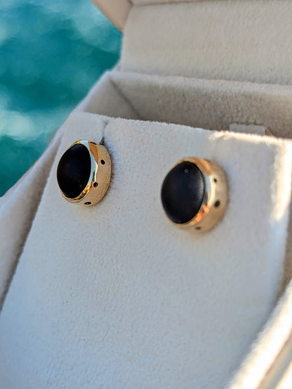 Pair of rare opaque black sea glass earrings set in solid 9ct yellow gold, displayed in a jewellery box with the ocean behind.