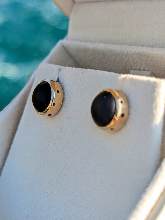 Pair of rare opaque black sea glass earrings set in solid 9ct yellow gold, displayed in a jewellery box with the ocean behind.