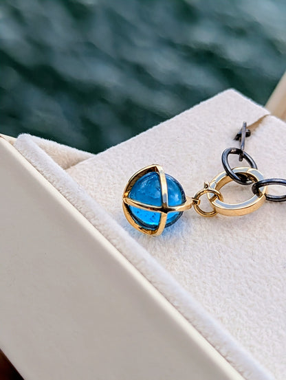Swiss Blue Topaz sphere pendant with a solid 9ct gold hinged charm holder beside a black rhodium sterling silver chain in an open jewellery box with ocean background.
