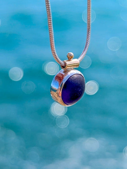 Detail of blue round sea glass silver necklace with gold bail detail above shimmering ocean background.
