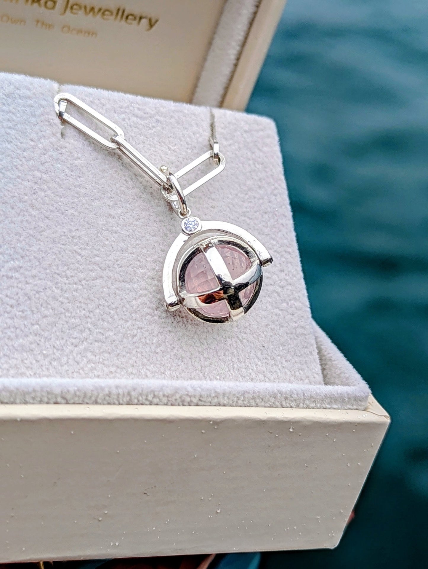 Silver ORB necklace with a pale pink rose quartz sphere and small moissanite, displayed in a jewellery box beside the sea.