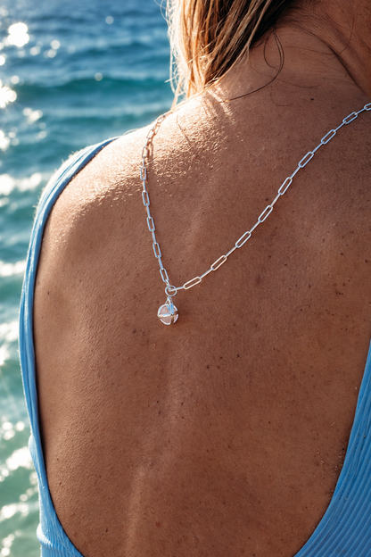 Woman in a blue swimsuit wearing a silver ORB spinning gemstone necklace down her bare back, with the sea behind her.