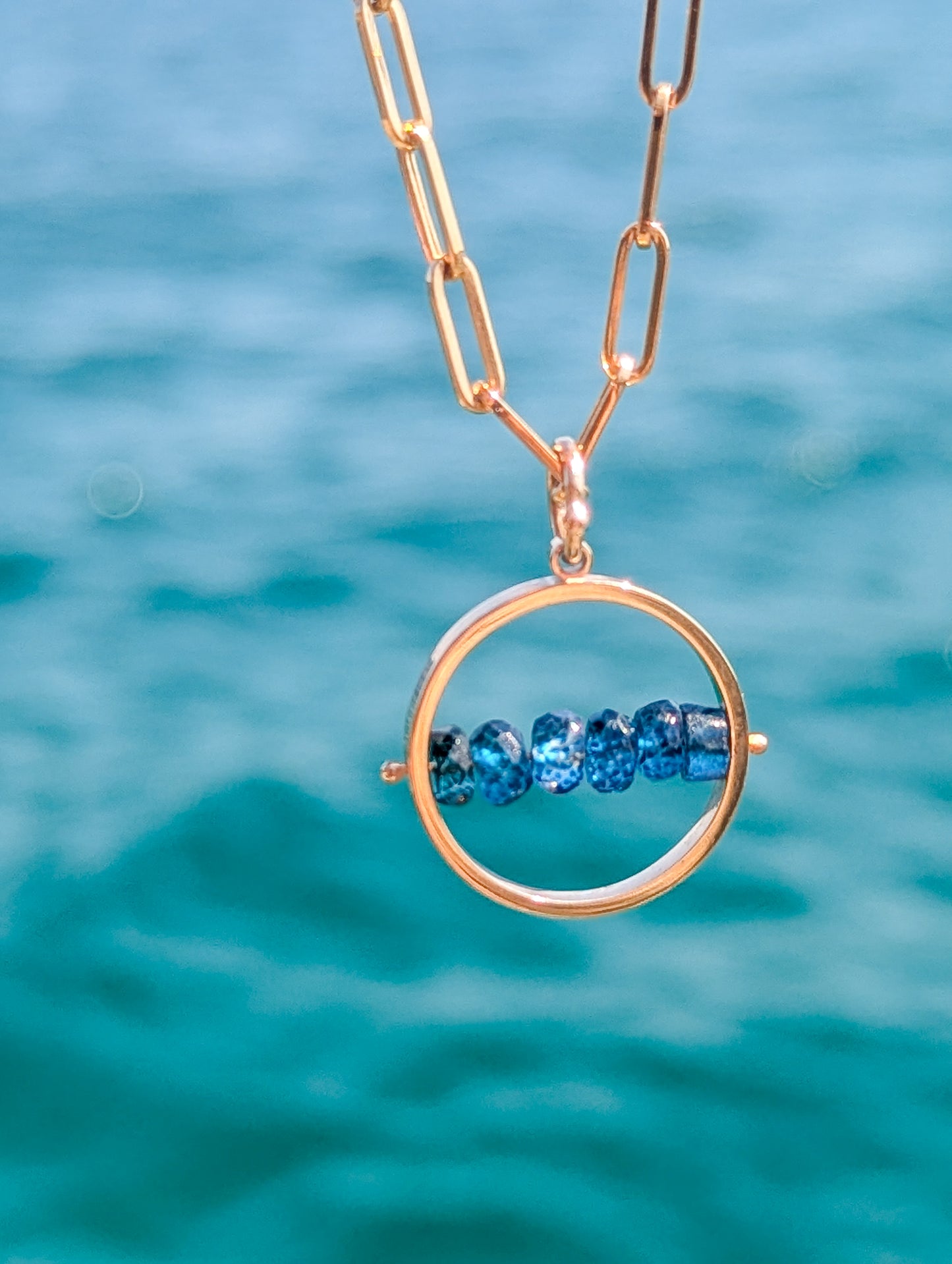 HALO solid 9ct gold spinning necklace with six natural London Blue Topaz gemstones against the ocean.