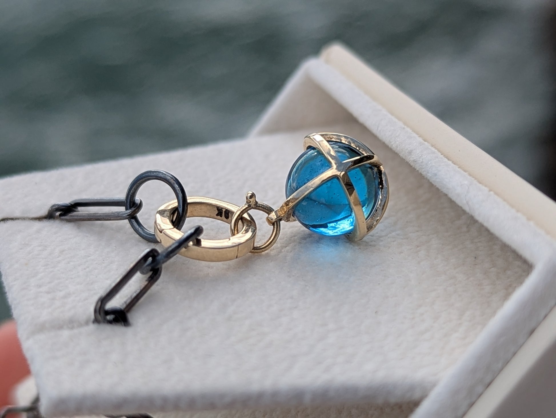 Swiss Blue Topaz sphere pendant with a solid 9ct gold hinged charm holder resting on coastal rocks beside a black rhodium sterling silver chain.