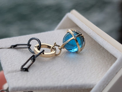 Swiss Blue Topaz sphere pendant with a solid 9ct gold hinged charm holder resting on coastal rocks beside a black rhodium sterling silver chain.